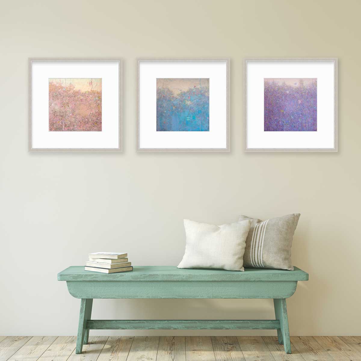 Pink, blue and dark pink abstract landscape prints in silver frames with mats hang on a beige wall above a light green bench with neutral pillows and a stack of books..