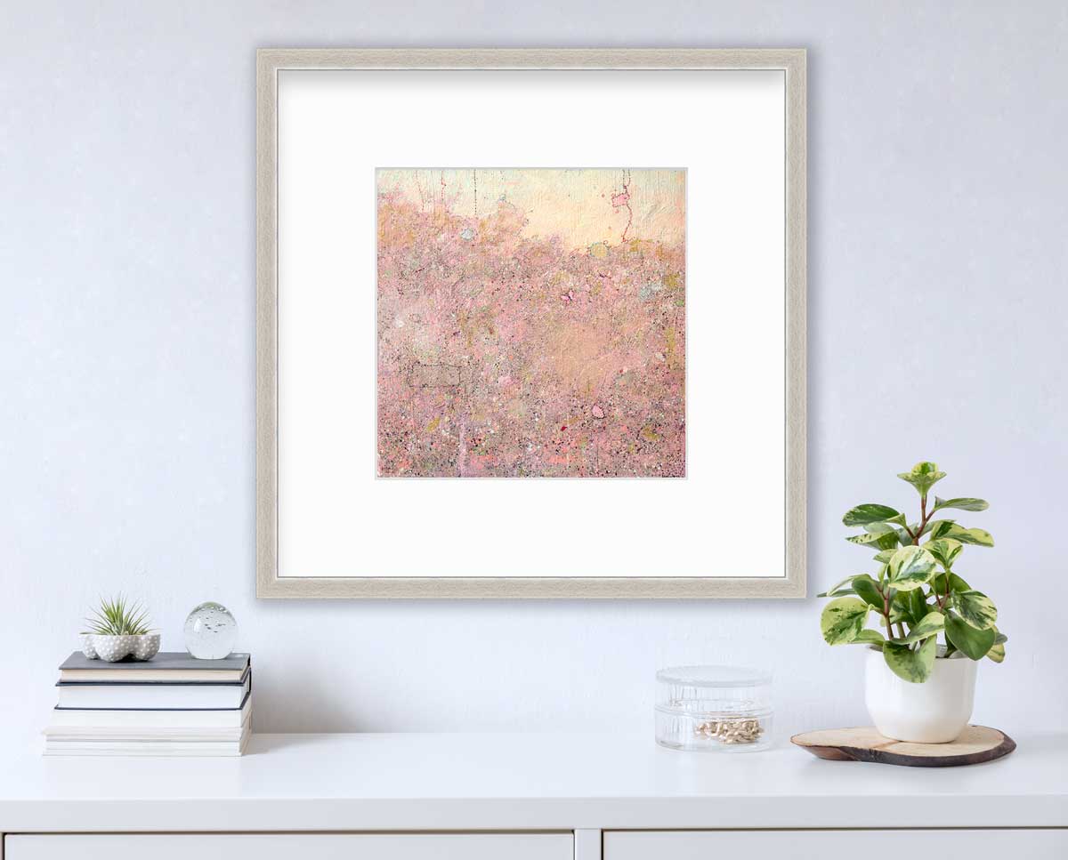 A pink abstract landscape print in a silver frame with a mat hangs on a white wall above a white surface with a stack of books, a clear trinket container with gold accessories and a potted plant.
