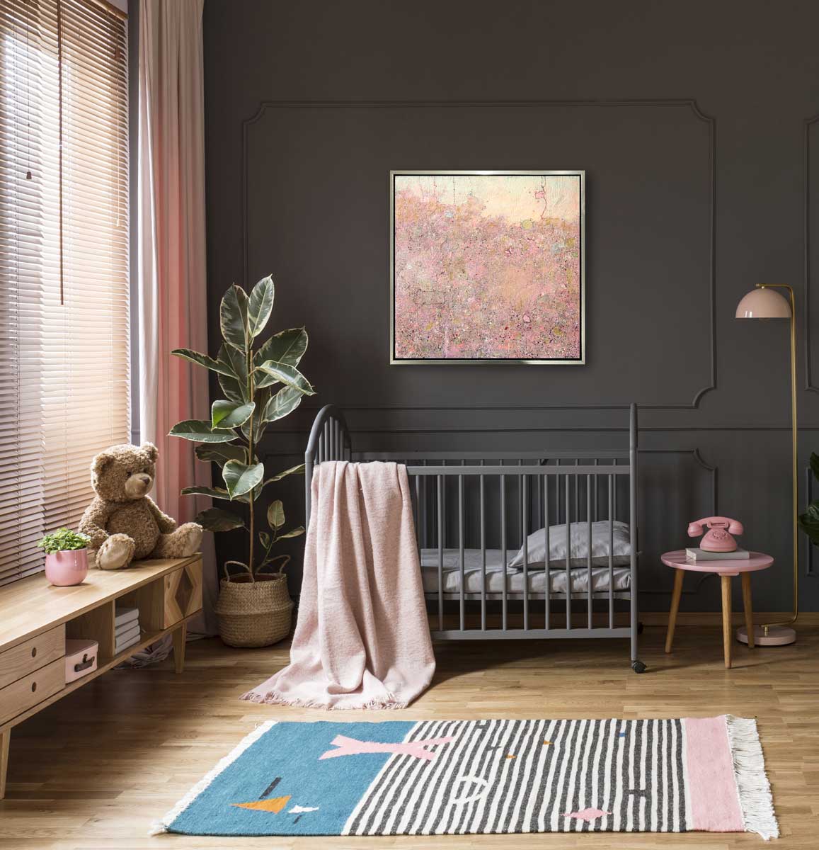 A pink abstract landscape print in a silver floater frame hangs on a dark grey paneled wall above a grey crib with a pink blanket hanging off the side, a large potted plant in the corner and a small side table with a pink rotary phone.