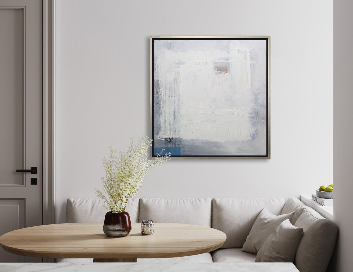 A white, grey and blue abstract print in a silver floater frame hangs on a white wall above a sofa like wrap around booth with white cushions and a natural wood table with a brown vase holding white floral stems.