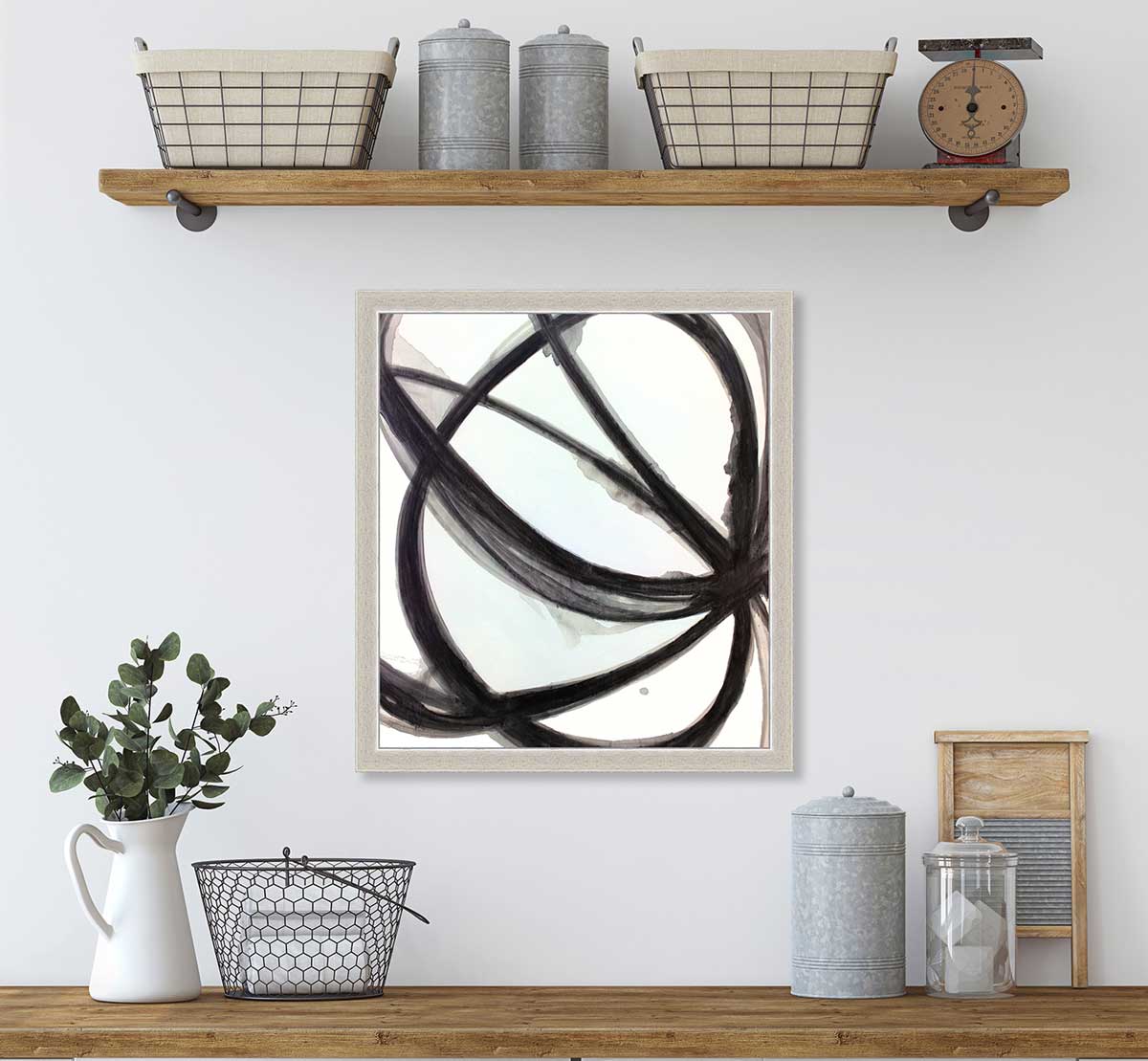 A black, grey and white abstract print in an unmatted silver frame hangs on a white wall above a butcher block countertop with a white vase filled with eucalyptus stems, a black wire basket and accessories.