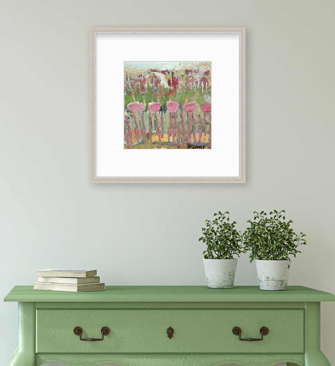 A green, pink and teal abstract floral print in a silver frame with a mat hangs on a white wall above a green console table with a stack of books and two potted plants.