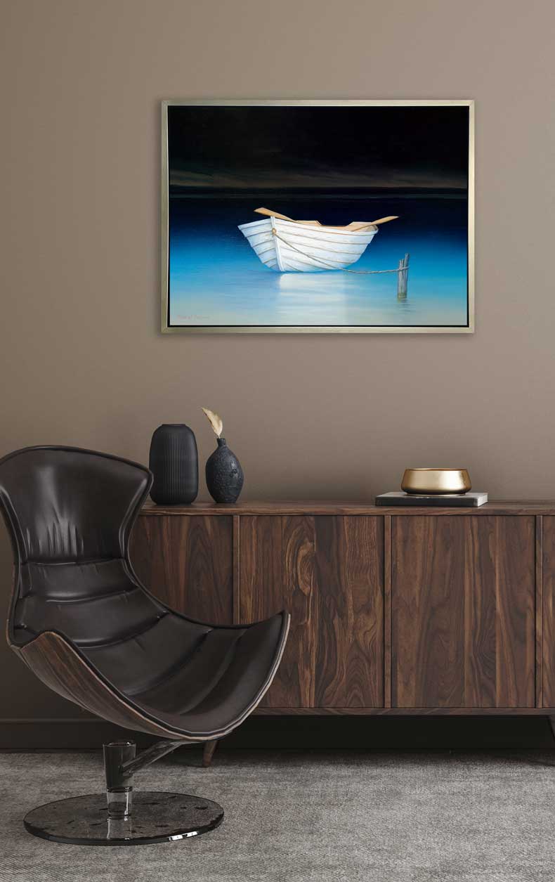 A contemporary realist print of a white rowboat alone on dark water against a dark sky in a silver floater frame hangs on a beige wall above a stained wood shelf with black vases and decorative items and a modern brown chair.
