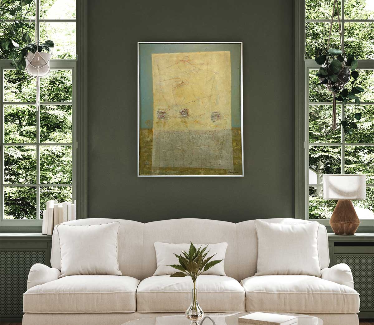 A teal, light yellow and grey abstract painting in a white floater frame hangs on a green wall in between two windows and above a white sofa with pillows.