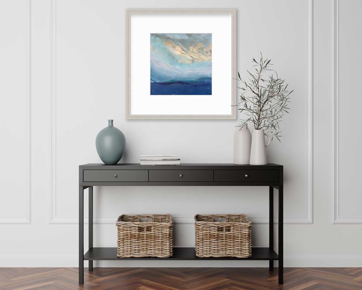 A blue, gold and teal abstract print in a silver frame with a mat hangs on a white wall above a black console table with a muted teal vase, a stack of books, two white vases with greenery stems and two wicker baskets.