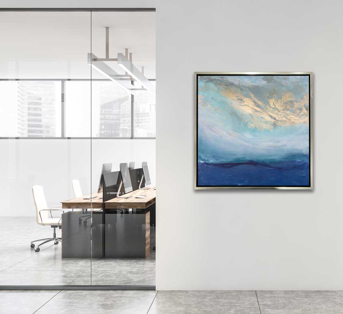 A blue, gold and teal abstract print in a silver floater frame hangs on a white wall next to two rows of desks with computers and desk chairs.