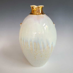 A white and cream glazed vessel with two small faux handles and a fluted gold opening sits on a white surface.