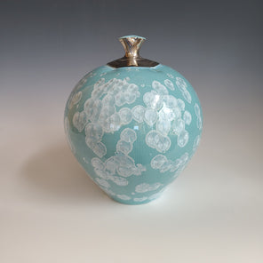 A light teal and white crystalized glazed vase with a small fluted opening sits on a white surface.