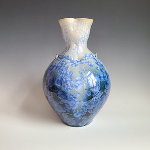 A cream, white and blue crystalized glazed narrow vase with two faux handles and a long fluted opening sits on a white surface.