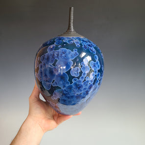 A hand holding a blue tonal crystalized vase with a charcoal long narrow opening.