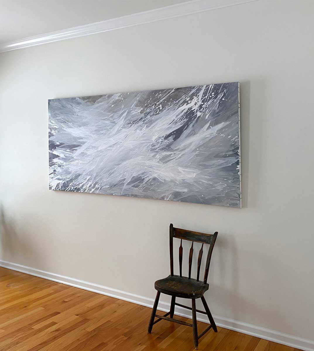 A white, lavender, and cool grey abstract painting by Teodora Guererra hangs on a white wall with a brown wood chair sitting below to the right and a potted plant to the left.