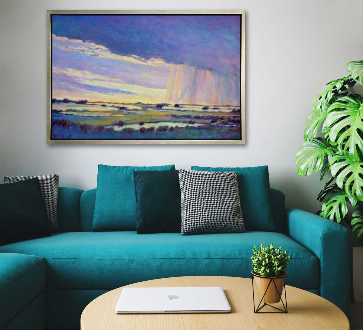A blue, purple and yellow abstract landscape print in a silver floater frame hangs on a white wall above a teal sofa with pillows, a round wooden coffee table with a laptop and small potted plant and a large potted floor plant.