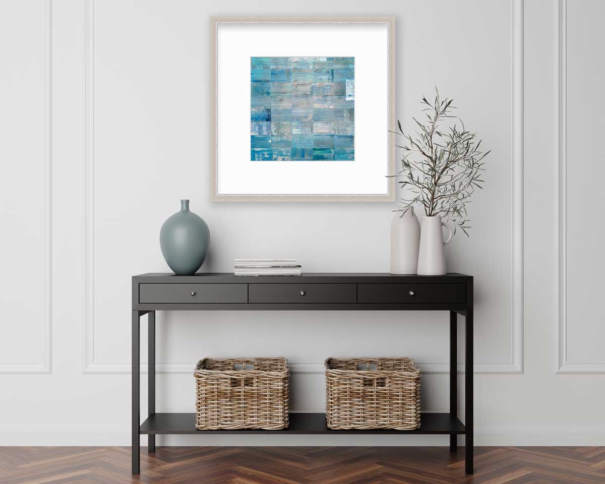 A blue and teal geometric print in a silver frame with a mat hangs on a white wall above a black console table with a muted teal vase, two white vases with greenery stems and two wicker baskets.