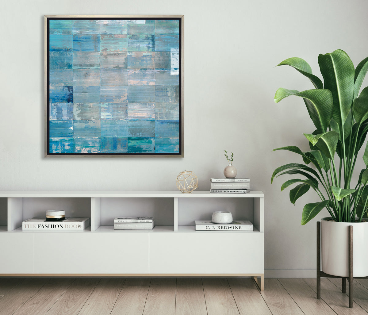 A blue and teal abstract geometric print in a silver floater frame hangs on a white wall above a white console cabinet with stacks of books, a gold wire sphere and a potted floor plant to one side.
