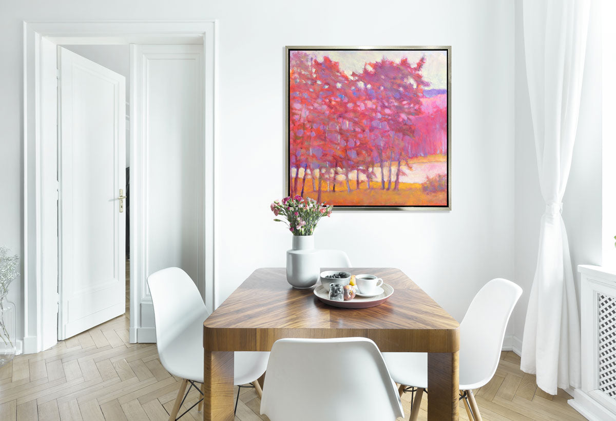 A red and pink tree landscape print in a silver floater frame hangs on a white wall above a wooden table and four white chairs.