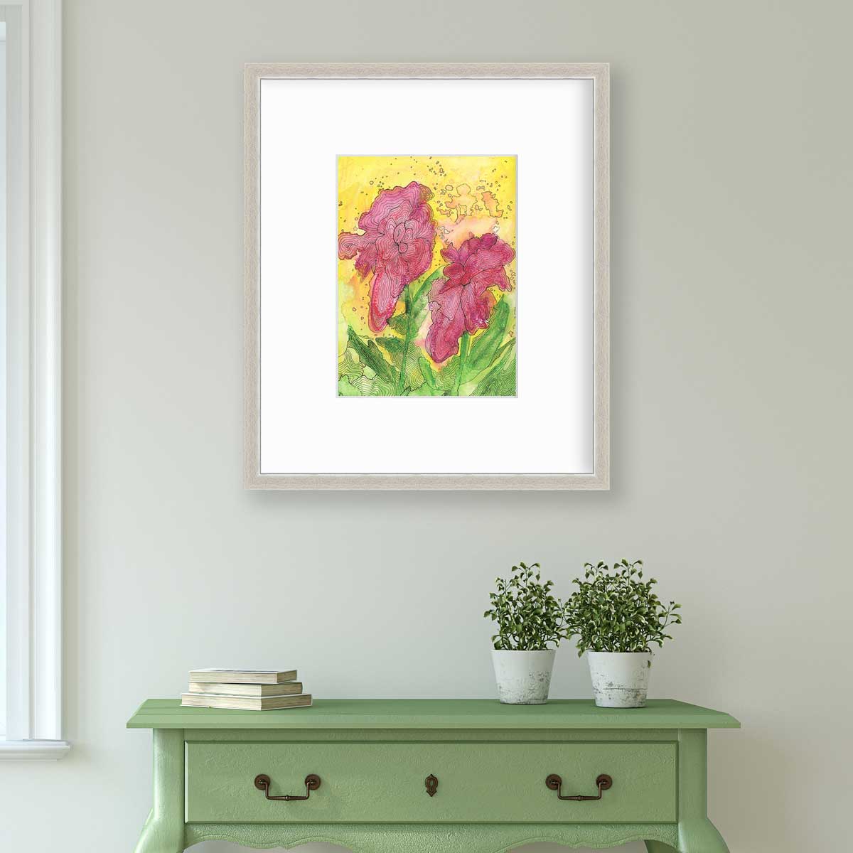 A red, yellow, green and black abstract floral print in a silver frame with a mat hangs on a white wall above a green console table with a stack of books and two potted plants.