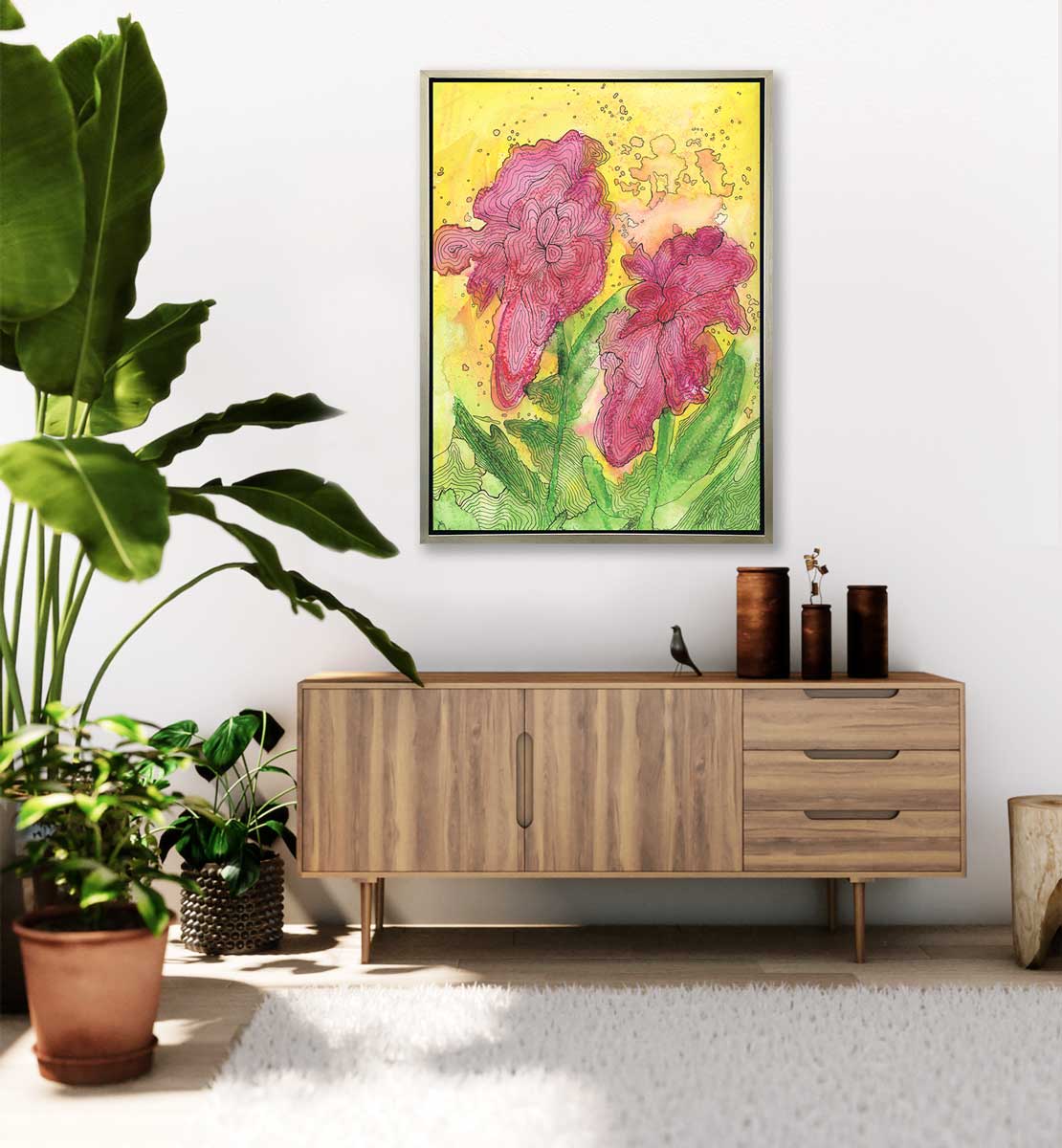 A red, yellow, green and black abstract floral print in a silver floater frame hangs on a white wall above a wood sideboard with three brown vases, a small bird figurine and several potted floor plants.
