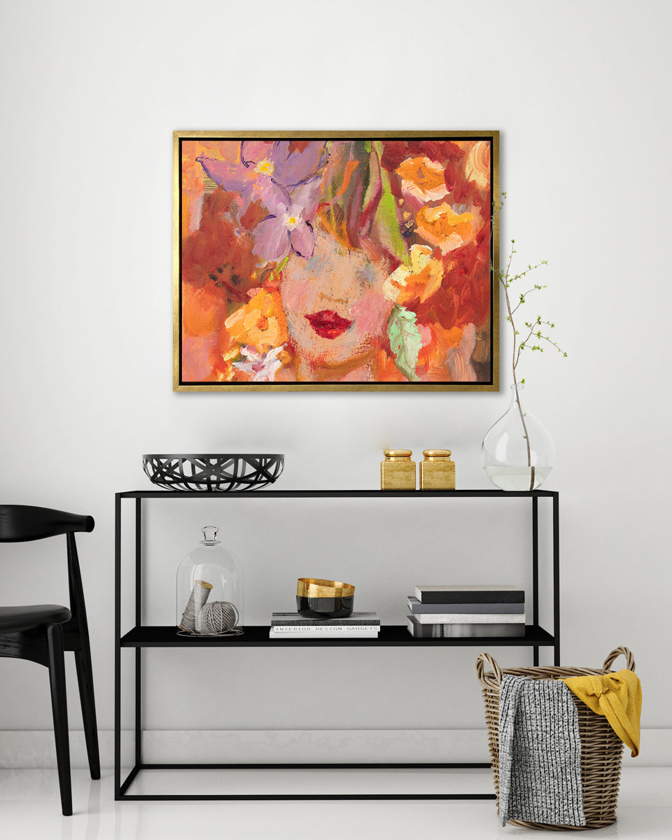 A red, orange, yellow and purple figural print in a gold floater frame hangs on a white wall above an open black frame shelf with a black wire bowl, two gold containers, stacks of books, a glass vase with a greenery stem and a woven basket on the floor with blankets.