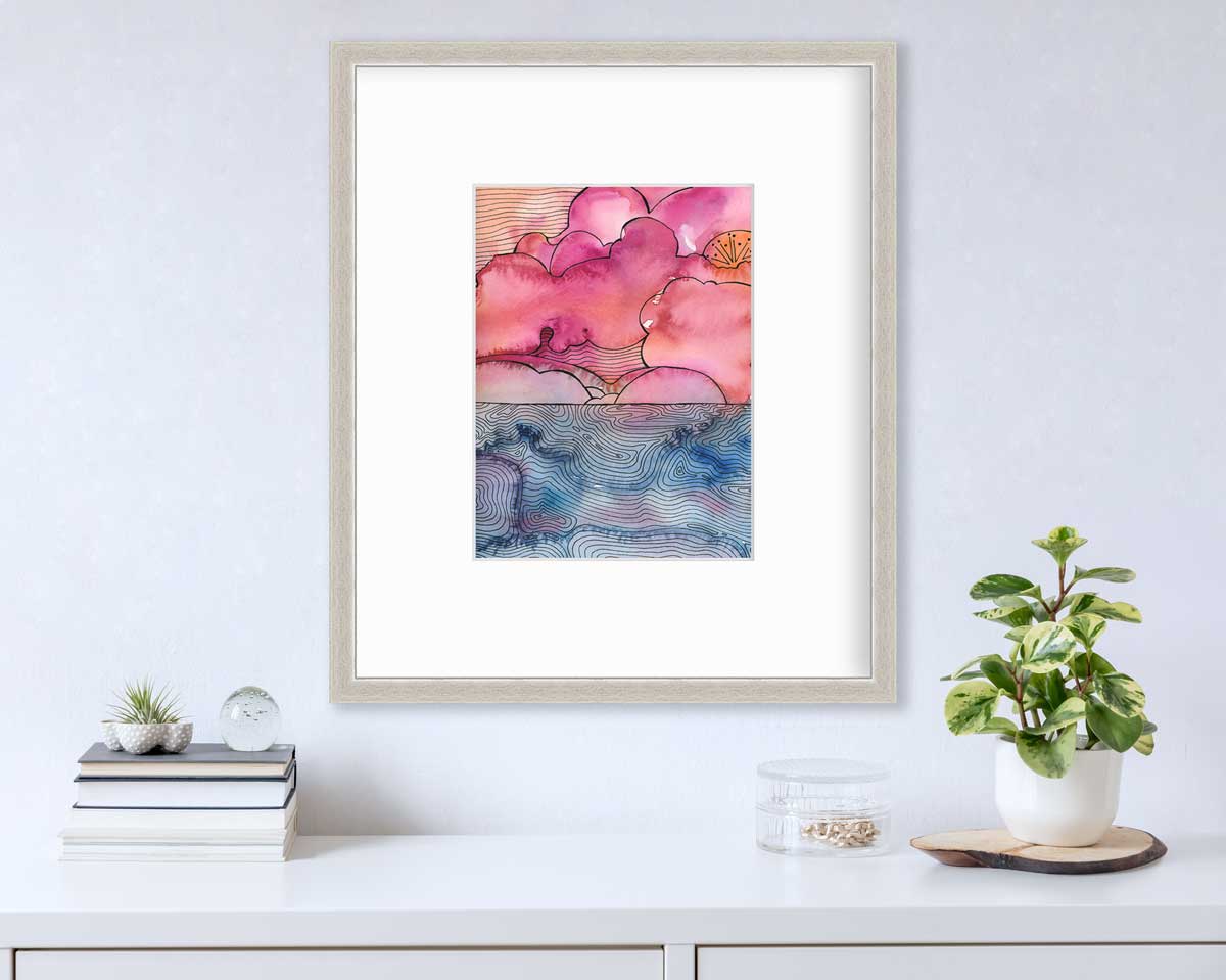 A pink, blue and black abstract landscape print in a silver frame with a mat hangs on a white wall above a white surface with a stack of books, a clear round container with gold accessories and a potted plant.
