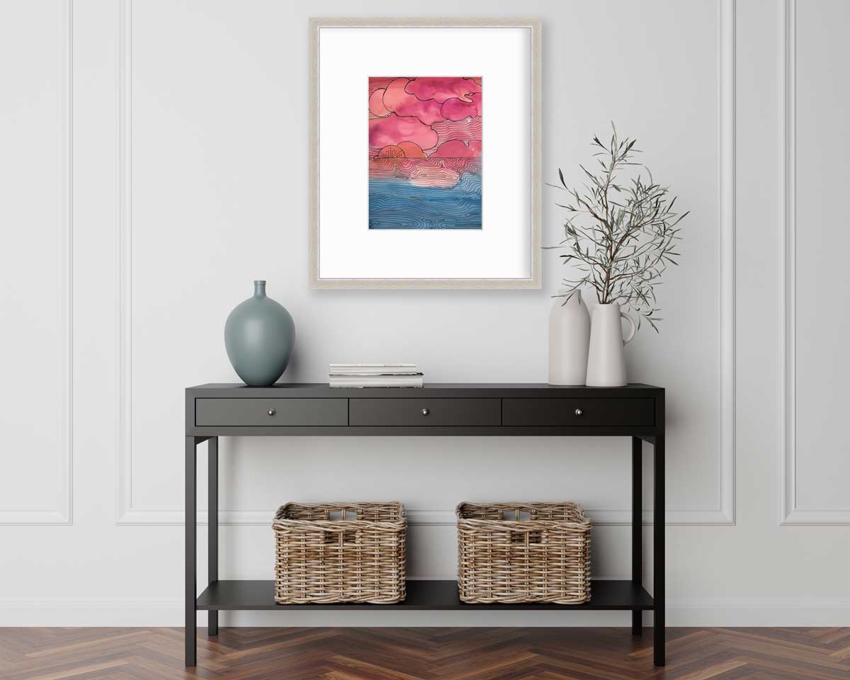 A pink, blue and black abstract landscape print in a silver frame with a mat hangs on a white wall above a black console table with a muted teal vase, a stack of books, two white vases with greenery stems and two wicker baskets,