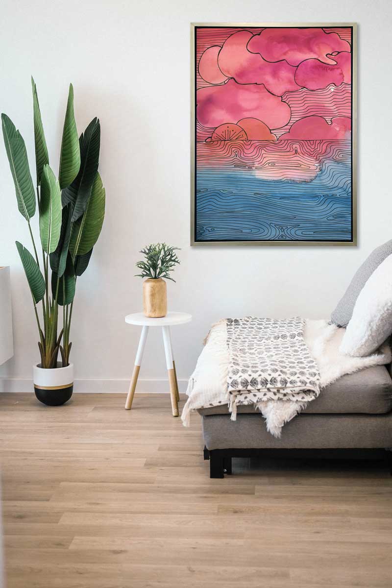 A pink, blue and black abstract landscape print in a silver floater frame hangs on a white wall above a grey loveseat with pillows and a white throw blanket, a small white side table with a gold vase with greenery and a potted floor plant.