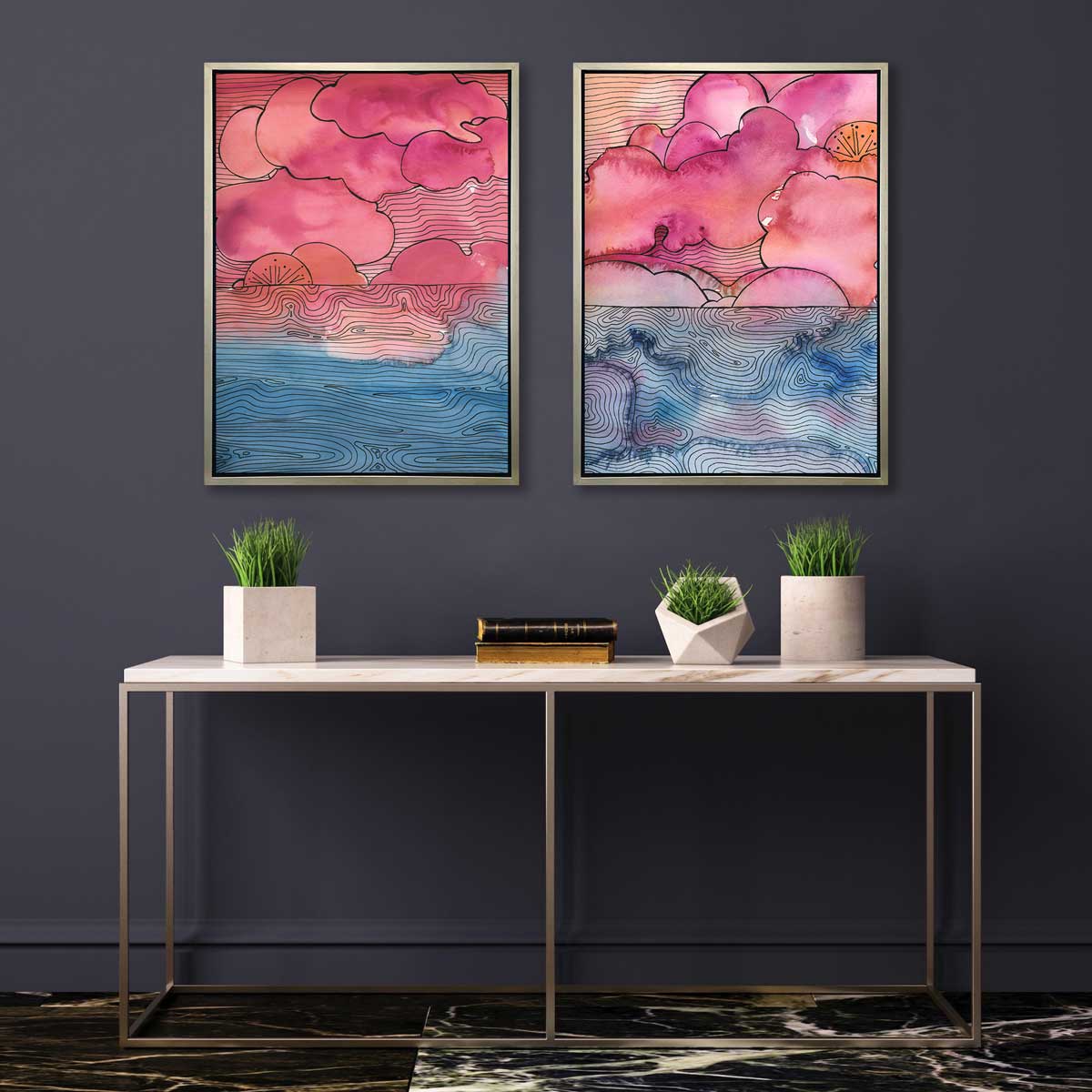 Two pink, blue and black abstract landscape prints in silver floater frames hang on a charcoal wall above a white marble console table with three greenery vases and two stacked books.