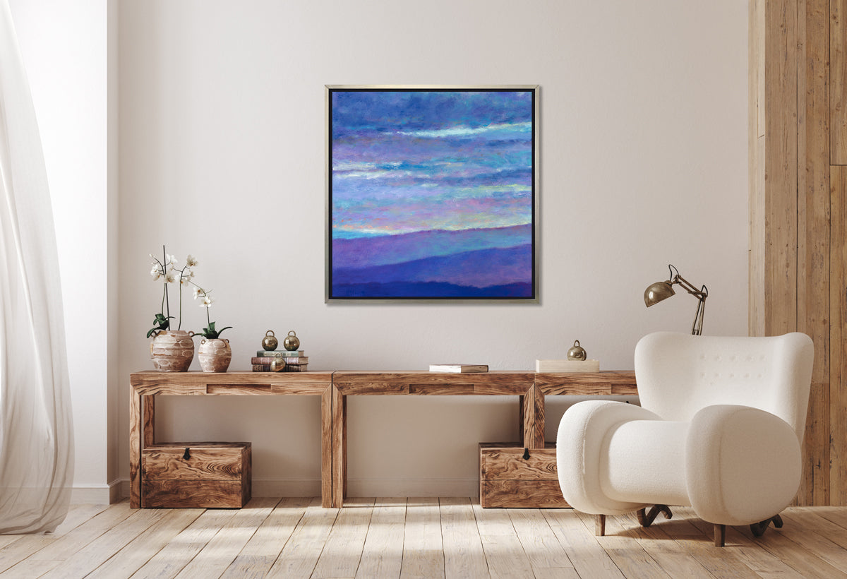 A blue, purple and light yellow abstract landscape print in a silver floater frame hangs on a white wall above a long natural wood desk with two potted white orchids, books and modern white armchair.