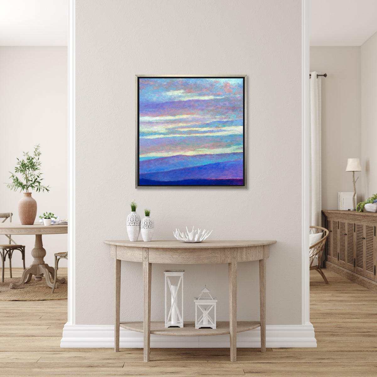 A blue, purple and light yellow abstract landscape print in a silver floater frame hangs on a white wall above a natural wood half-circle console table with two white vases, a white decorative bowl and two white lanterns.