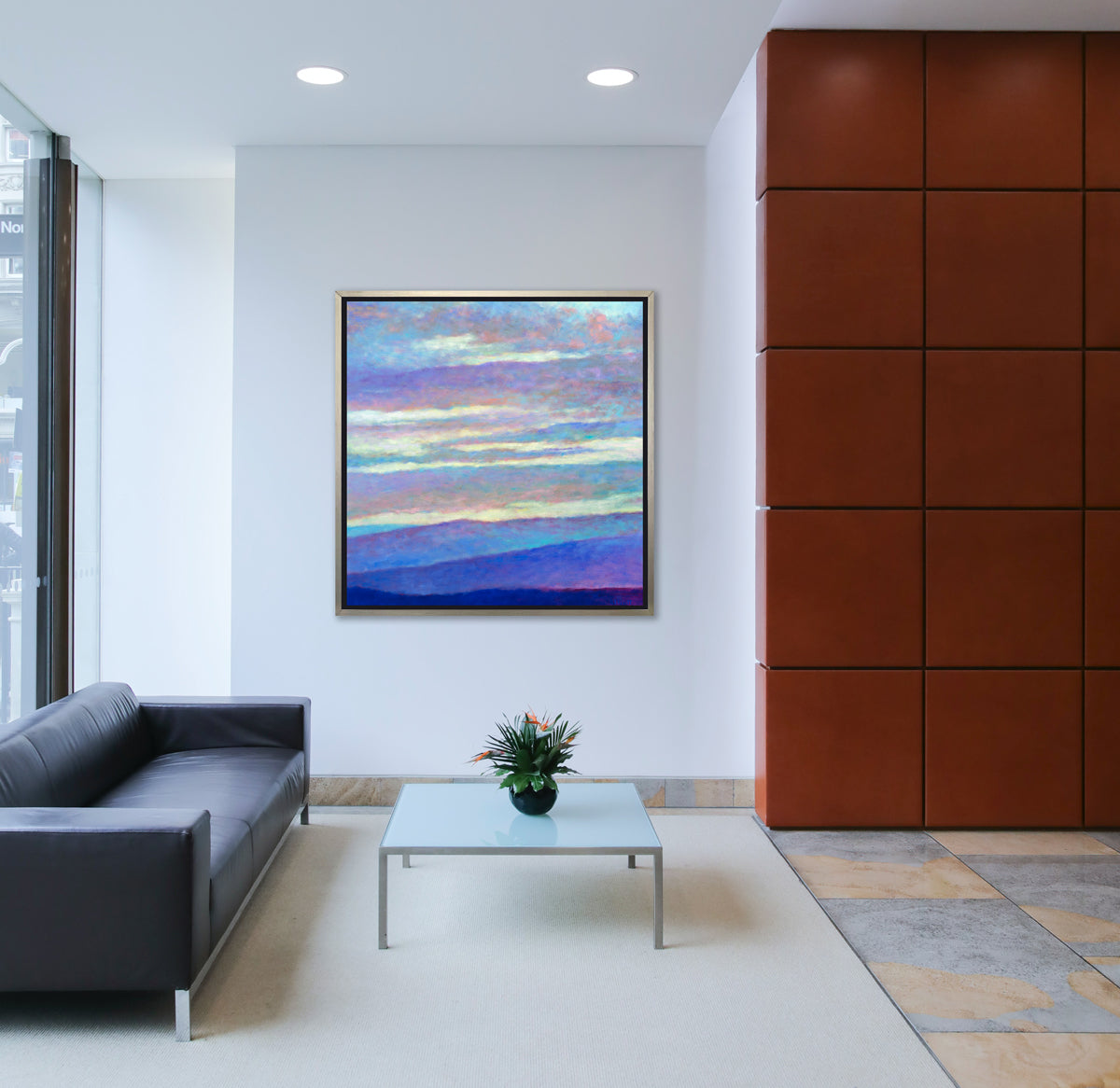 A blue, purple and light yellow abstract landscape print in a silver floater frame hangs on a white wall above a black leather sofa and a square frosted glass coffee table with a floral arrangement.