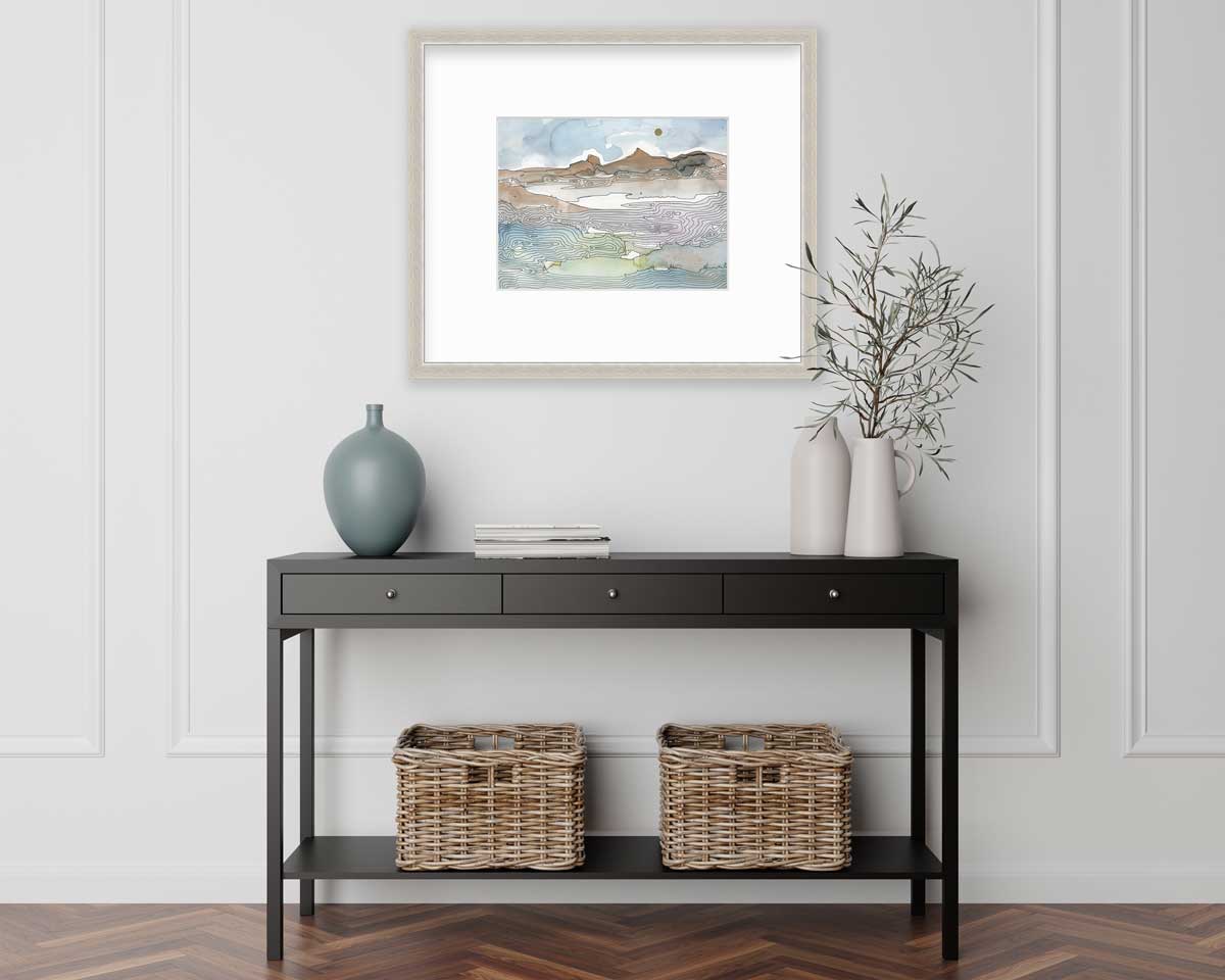 A blue, brown and light green abstract landscape print in a silver frame with a mat hangs on a white wall above a black console table with a muted teal vase, two white vases with greenery stems and two wicker baskets.