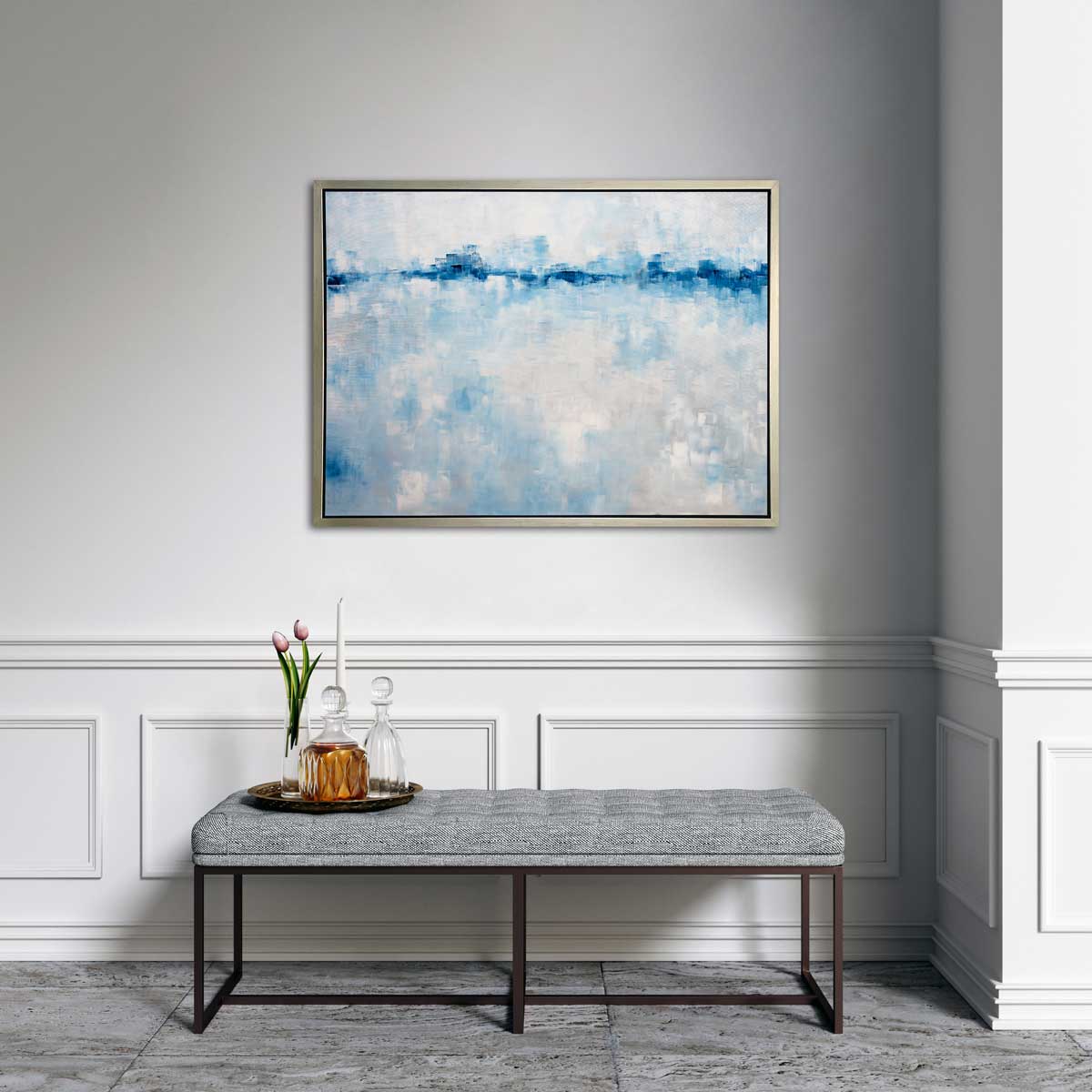 A blue and white abstract print in a silver floater frame hangs on a white wall above a grey tufted bench with a tray of two glass decanters, a white tapered candle and a clear vase with pink tulips.