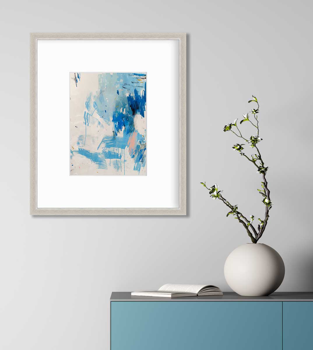 A blue, peach and white abstract print in a silver frame with a mat hangs on a white wall above a teal cabinet with an open book and a round white vase with floral stems.