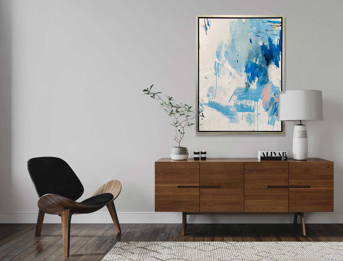 A blue, peach and white abstract print in a silver floater frame hangs on a white wall above a wood cabinet with a white lamp, a stack of books, a potted plant and a modern black chair off to the side.