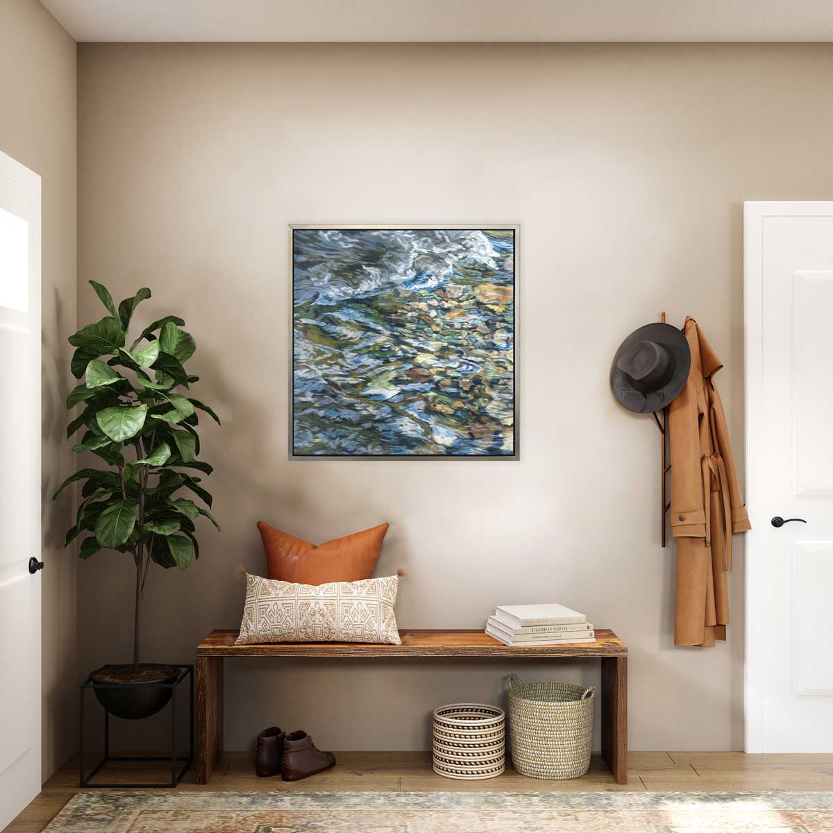 An abstract riverbed print in a silver floater frame hangs on a beige wall above a wooden bench with pillows and stacked books, a pair of brown shoes and two woven baskets.