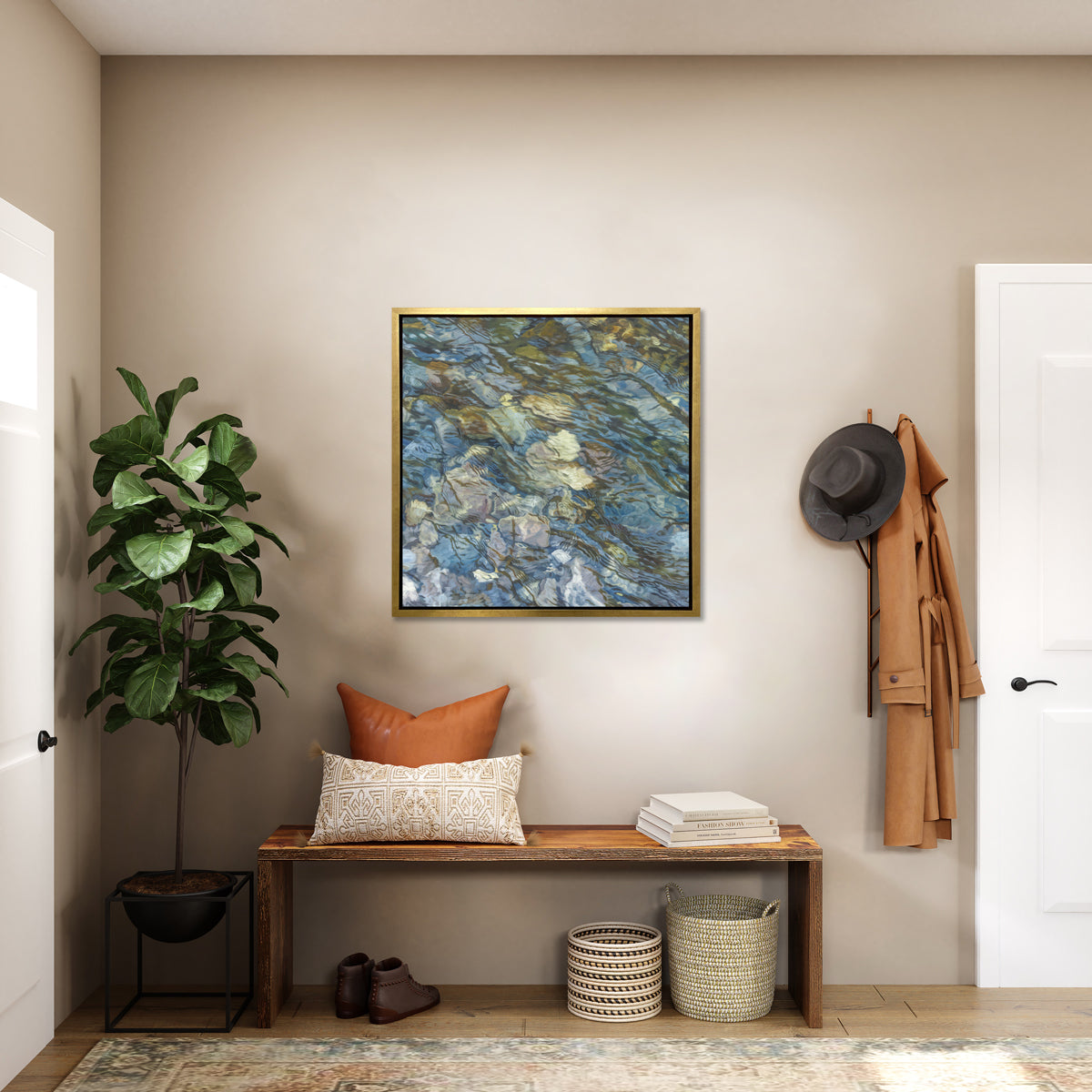 An abstract river bed print in a gold floater frame hangs on a white wall above a natural wood bench with pillows and a stack of books, two woven baskets, a pair of brown shoes and a tall potted plant.
