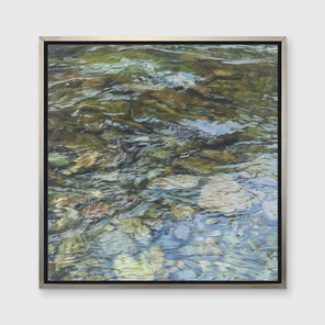 A green, brown, beige and white abstract river rock in water print in a silver floater frame hangs on a white wall.