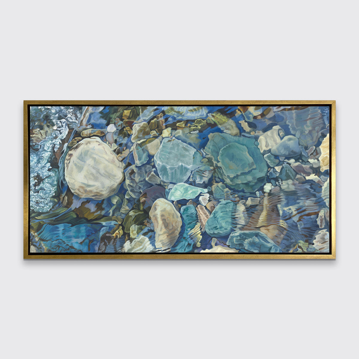 Abstracted print of grey, brown, green and beige river rocks under water hangs in a gold floater frame on a white wall.