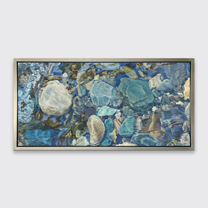 Abstracted print of grey, brown, green and beige river rocks under water hangs in a silver floater frame on a white wall.