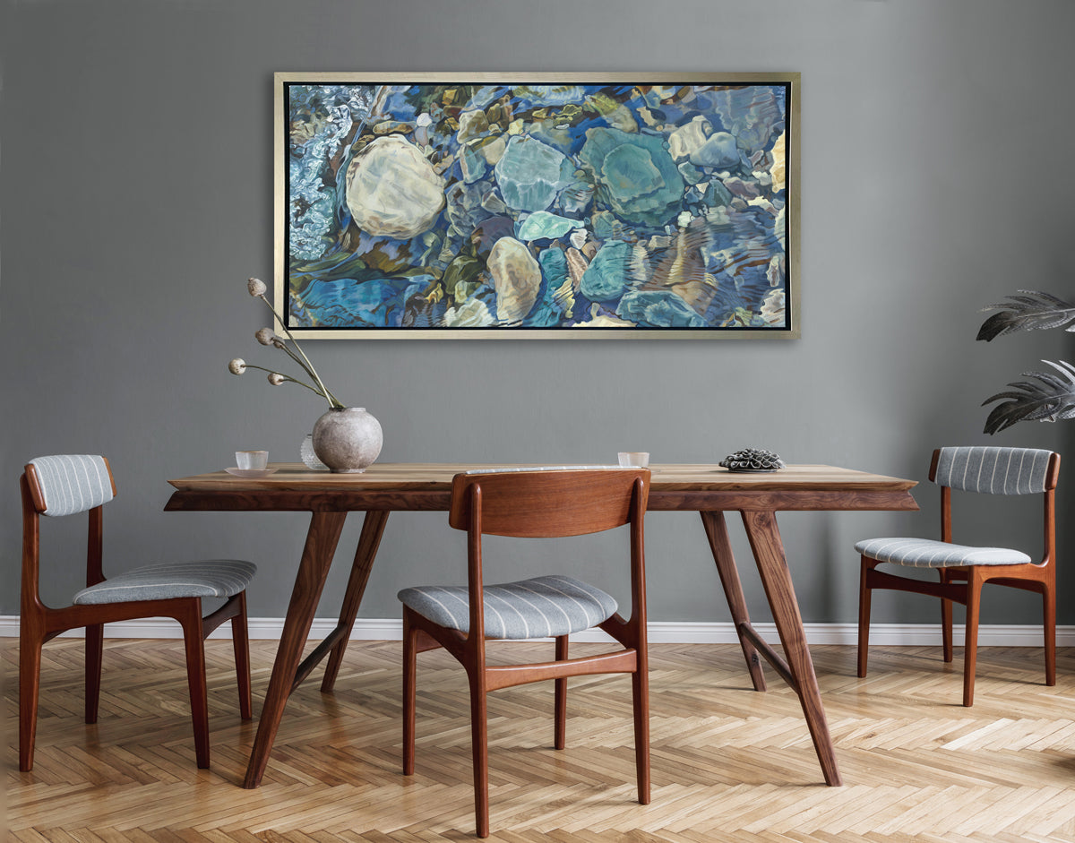 Abstracted print of grey, brown, green and beige river rocks under water hangs in a silver floater frame on a grey wall above a wood table with a floral filled vase, two small cups and three grey, white and stained wood chairs.
