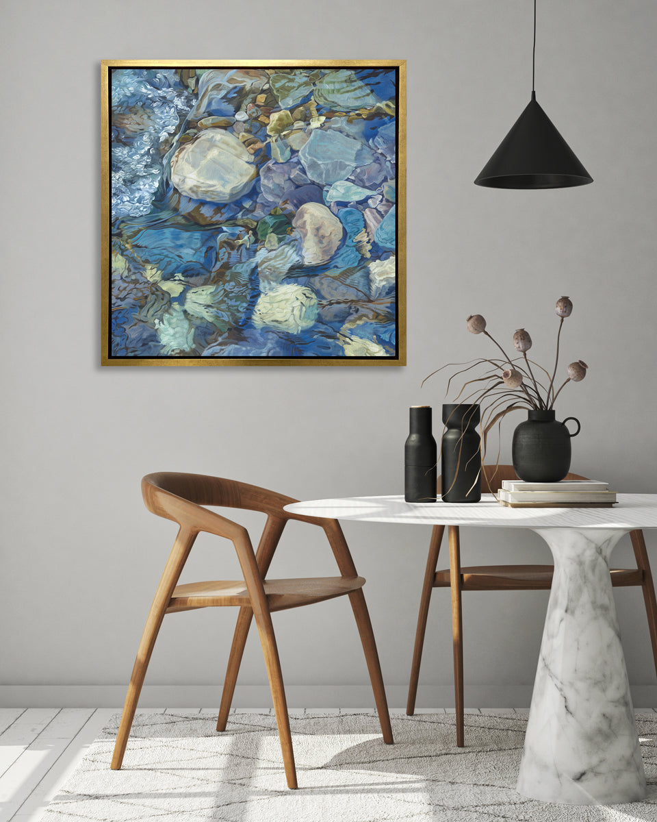 Abstracted print of grey, brown, green and blue river rocks under water hangs in a gold floater frame on a white wall above a white marble pedestal table with black vases and two smooth natural wood modern chairs.