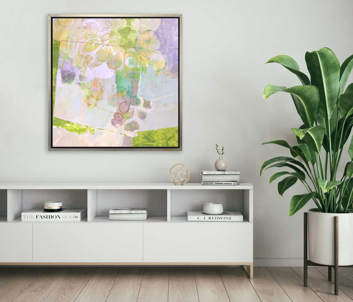 A light yellow, lavender and green abstract floral print in a silver floater frame hangs on a white wall above a white console shelf with books, a gold wire sphere and a potted floor plant.