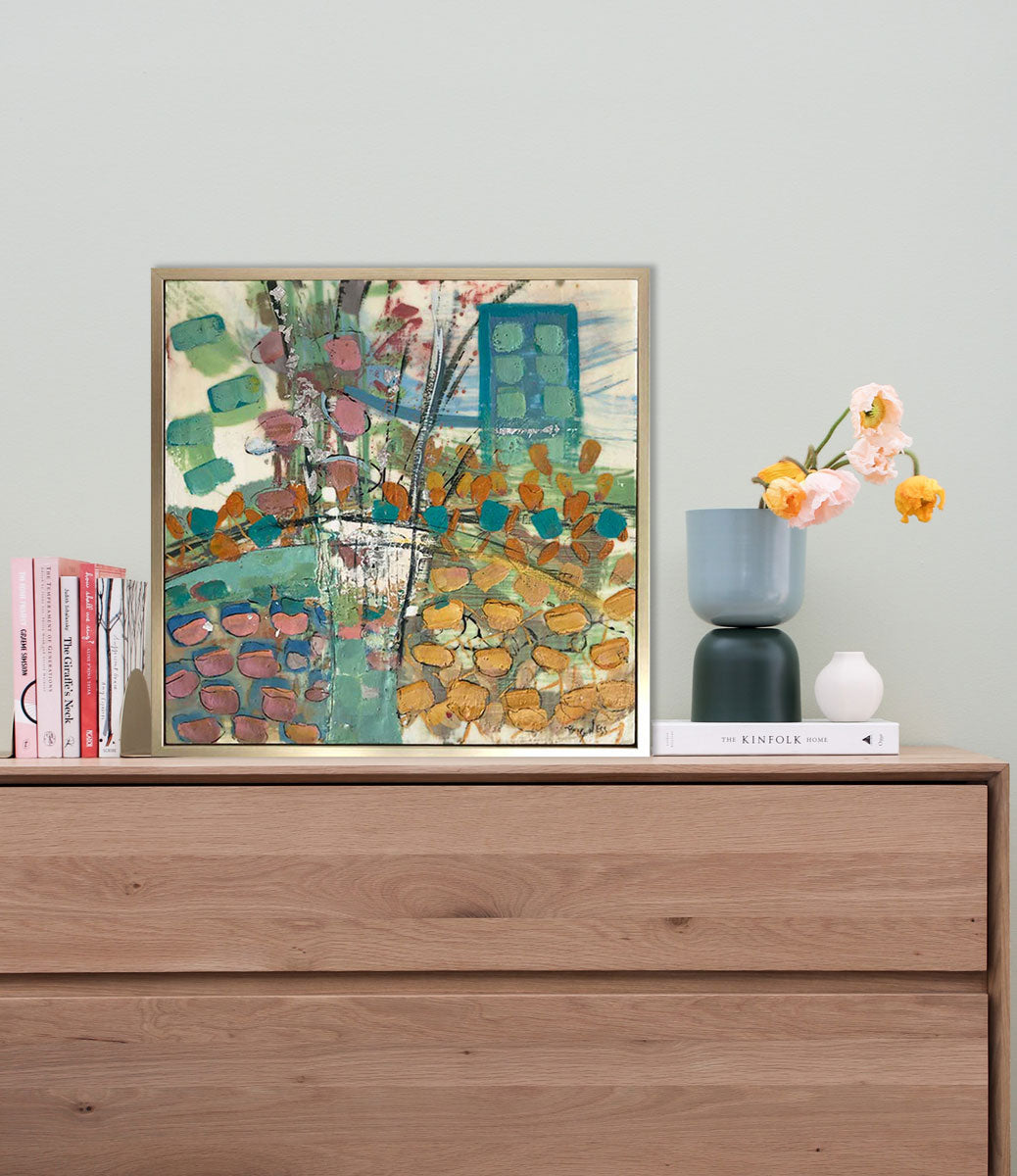 A multicolored abstract encaustic painting, framed in a gold frame, rests on top of a wooden dresser next to books and a modern vase with flowers. 