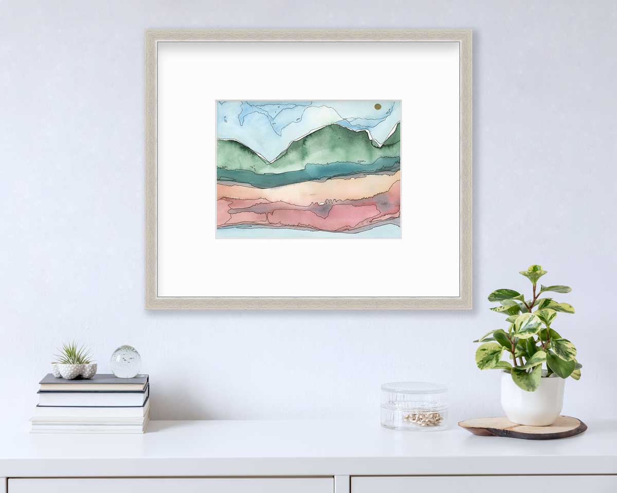 A blue, green and red abstract landscape print in a silver frame with a mat hangs on a white wall above a white surface with a stack of books, a clear round container with gold accessories and a potted plant.
