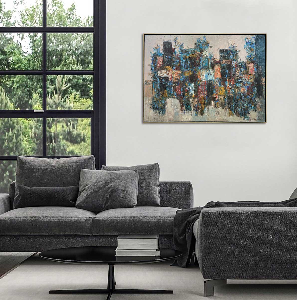 A multicolored abstract painting in a warm gold floater frame hangs on a white wall next to a large black paned window, above a grey sofa with pillows and a low round black coffee table.