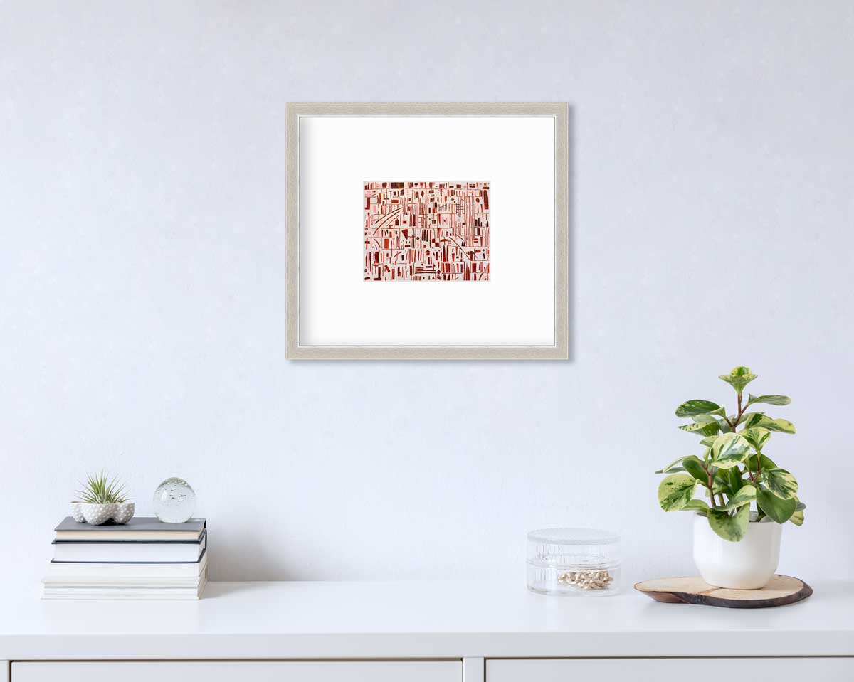 A red, pink and white abstract print in a silver frame with a mat hangs on a white wall above a white surface with a stack of books, a round clear container with gold accessories and a potted plant.