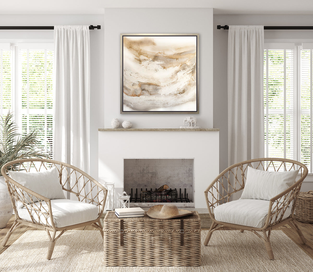 A gold, silver, white and beige abstract print in a silver floater frame hangs on a white wall above a white fireplace, two woven chairs with white cushions and a wicker trunk.