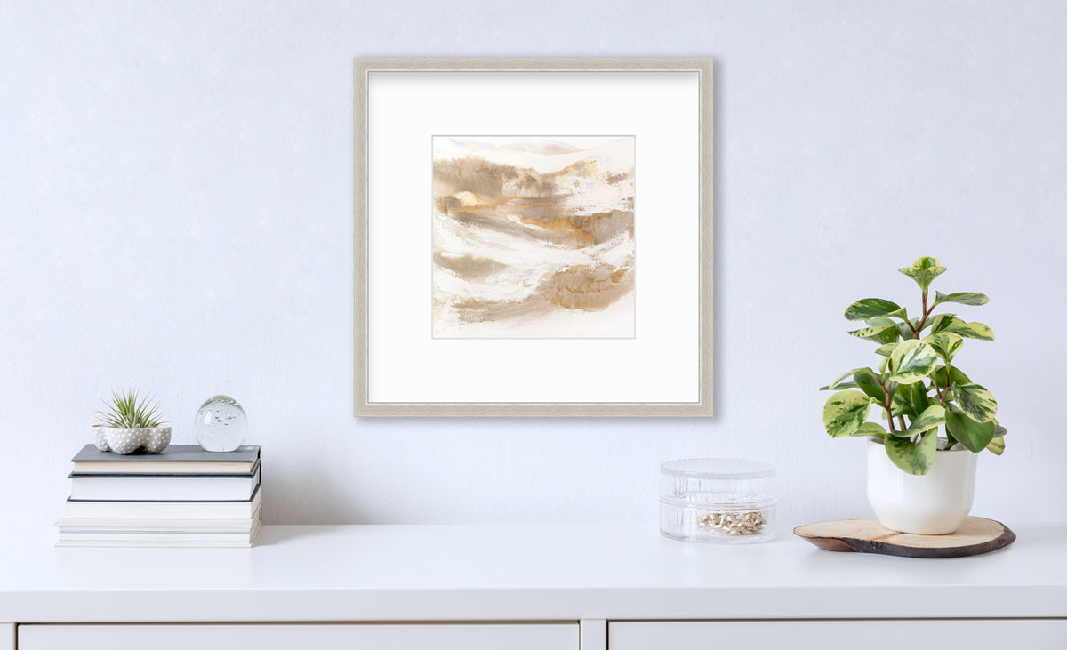 A gold, silver, white and beige abstract print in a silver frame with a mat hangs on a white wall above a white surface with a stack of books, a round clear container with gold accessories and a potted plant.