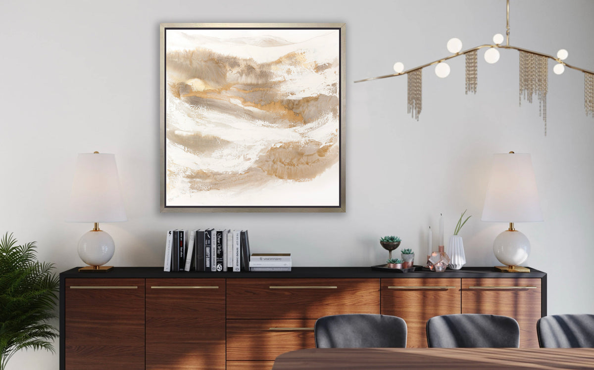 A gold, silver, white and beige abstract print in a silver floater frame hangs on a white wall above a black and dark wood sideboard with a stack of books, two matching white table lamps and small decorative accessories.