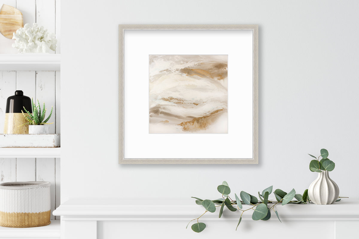 A gold, silver, white and beige abstract print in a silver frame with a mat hangs on a white wall above a white shelf with a eucalyptus filled white vase and next to a built in bookshelf with decorative accessories.