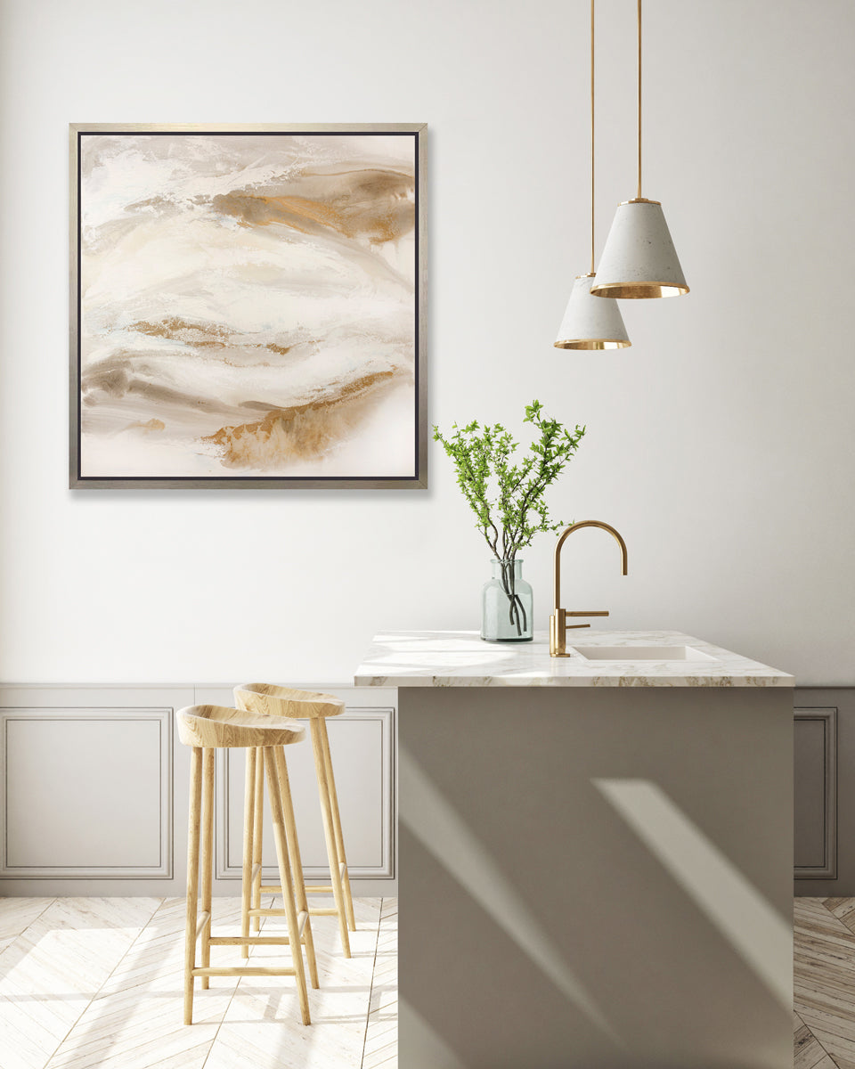 A gold, silver, white and beige abstract print in a silver floater frame hangs on a white wall above a white marble kitchen island with a sink and a glass bottle with greenery stems and two wood counter stools.